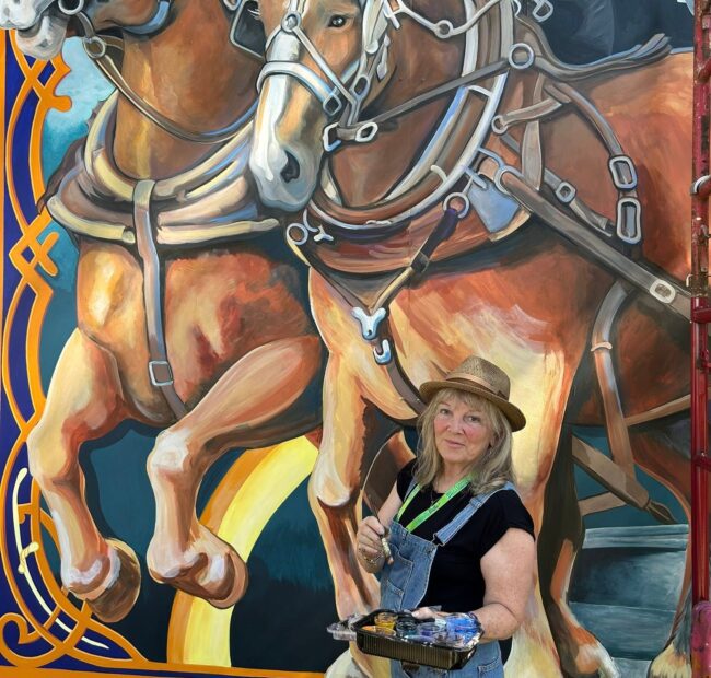 Woman with paint and brush in front of large mural of horses