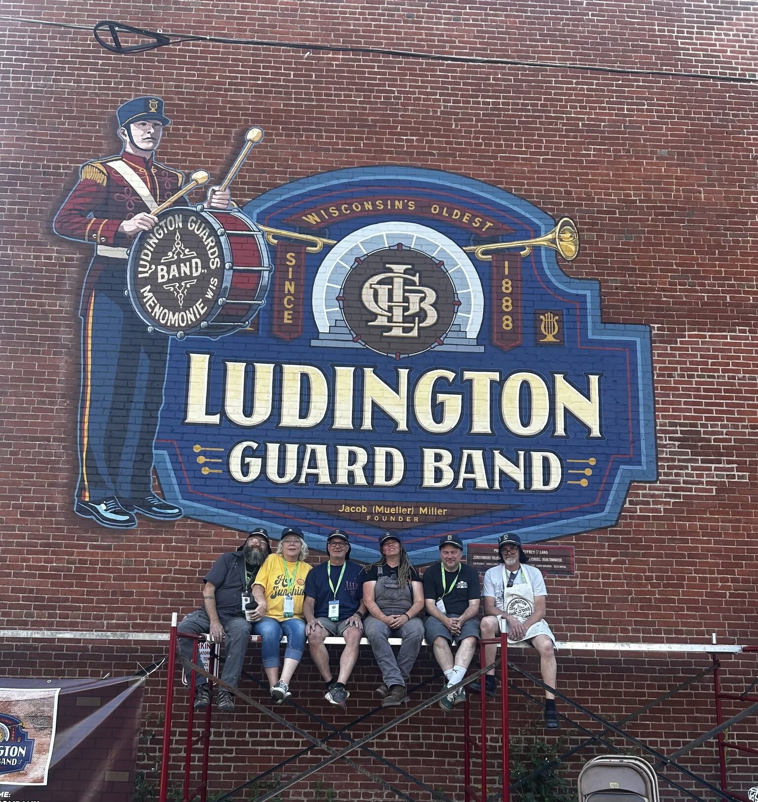 Group of people sitting on top of scaffolding in front of the Ludington Guard Band mural
