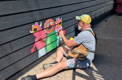 Man sitting on the ground painting a small mural of monsters on a black wall