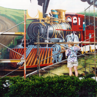 person under scaffolding painting a mural with large train