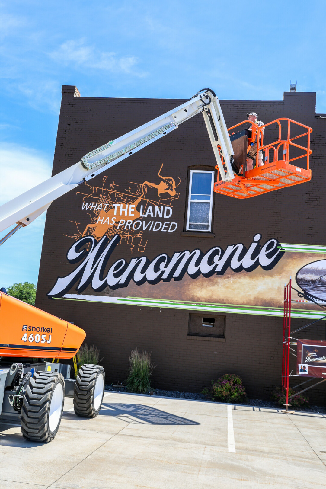 Man standing in a lift in front of a large mural that says What the land has provided, Menomonie