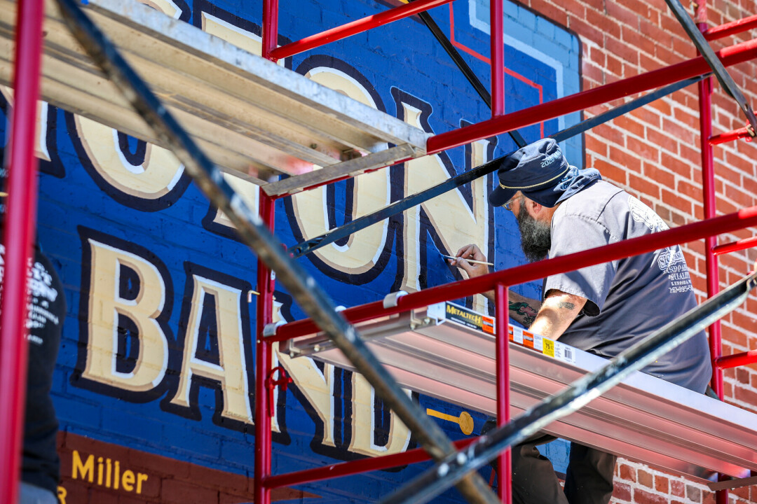 Man sitting on scaffolding painting a mural on a brick wall