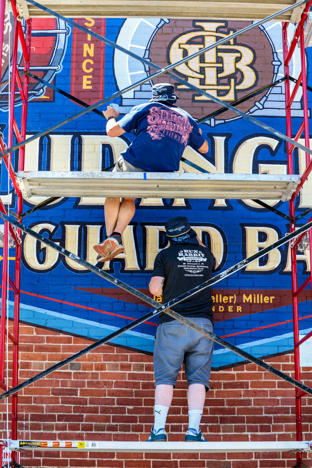 One person sitting above another person standing on scaffolding while painting on a brick wall