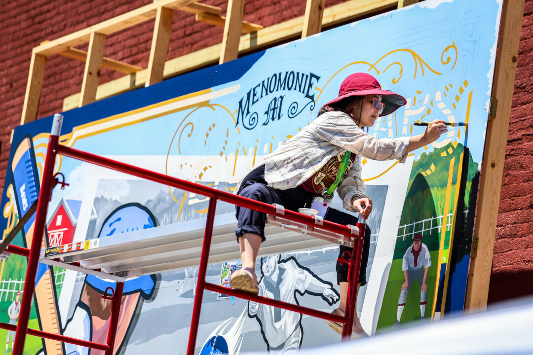 Lady in a hat painting a large mural while sitting on scaffolding.