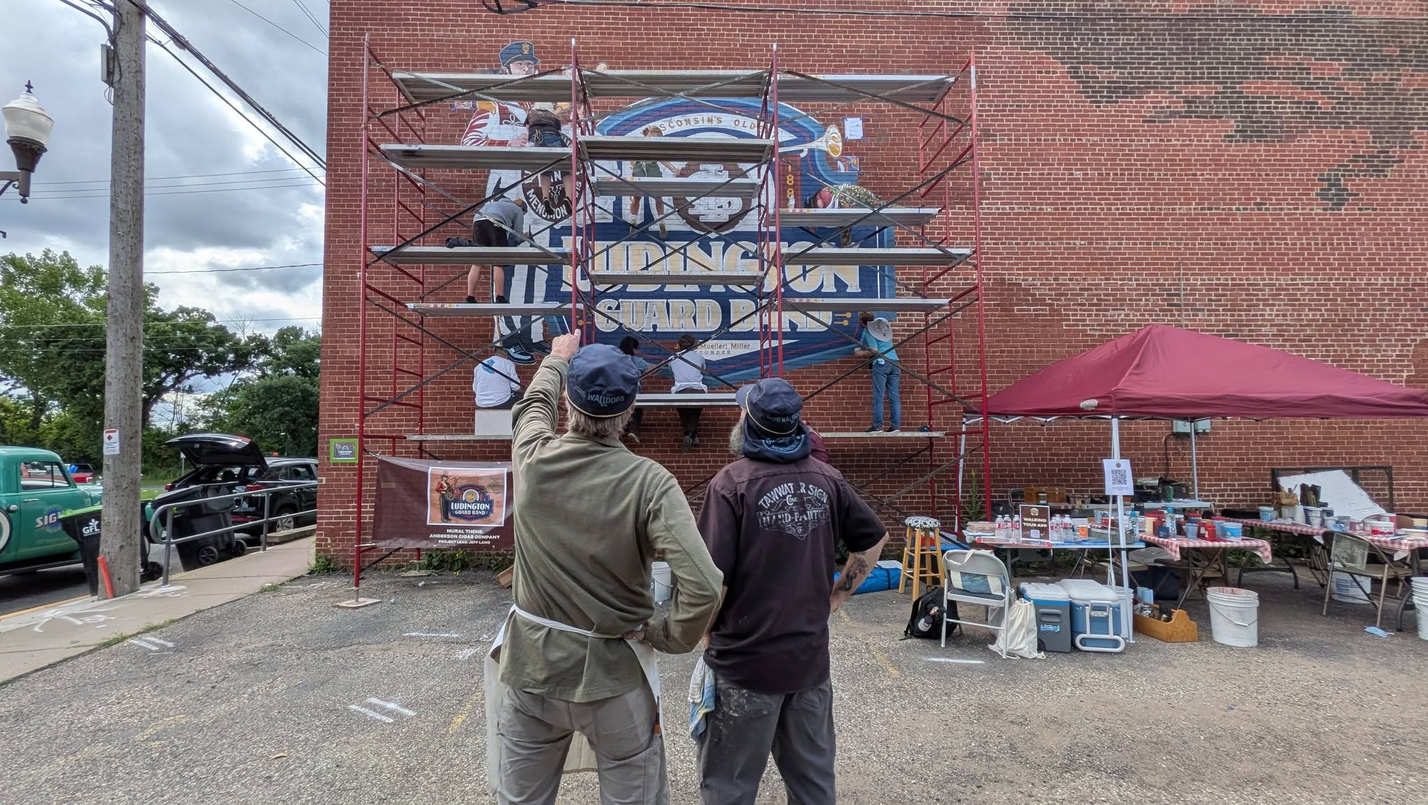 Backside of two people standing on the ground, one pointing at the mural with layers of scaffolding in front
