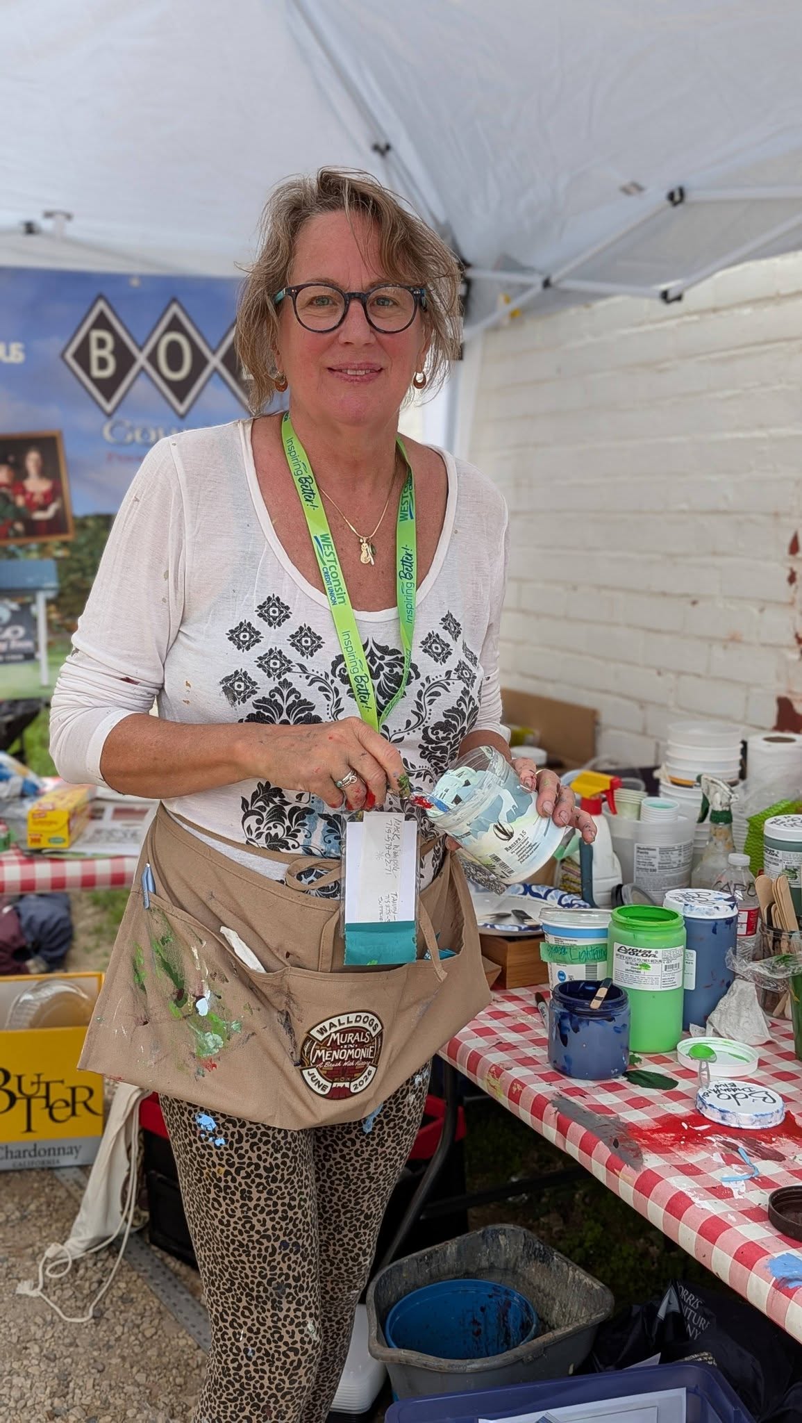 Artist in an apron holding paint by a table filled with paint canisters
