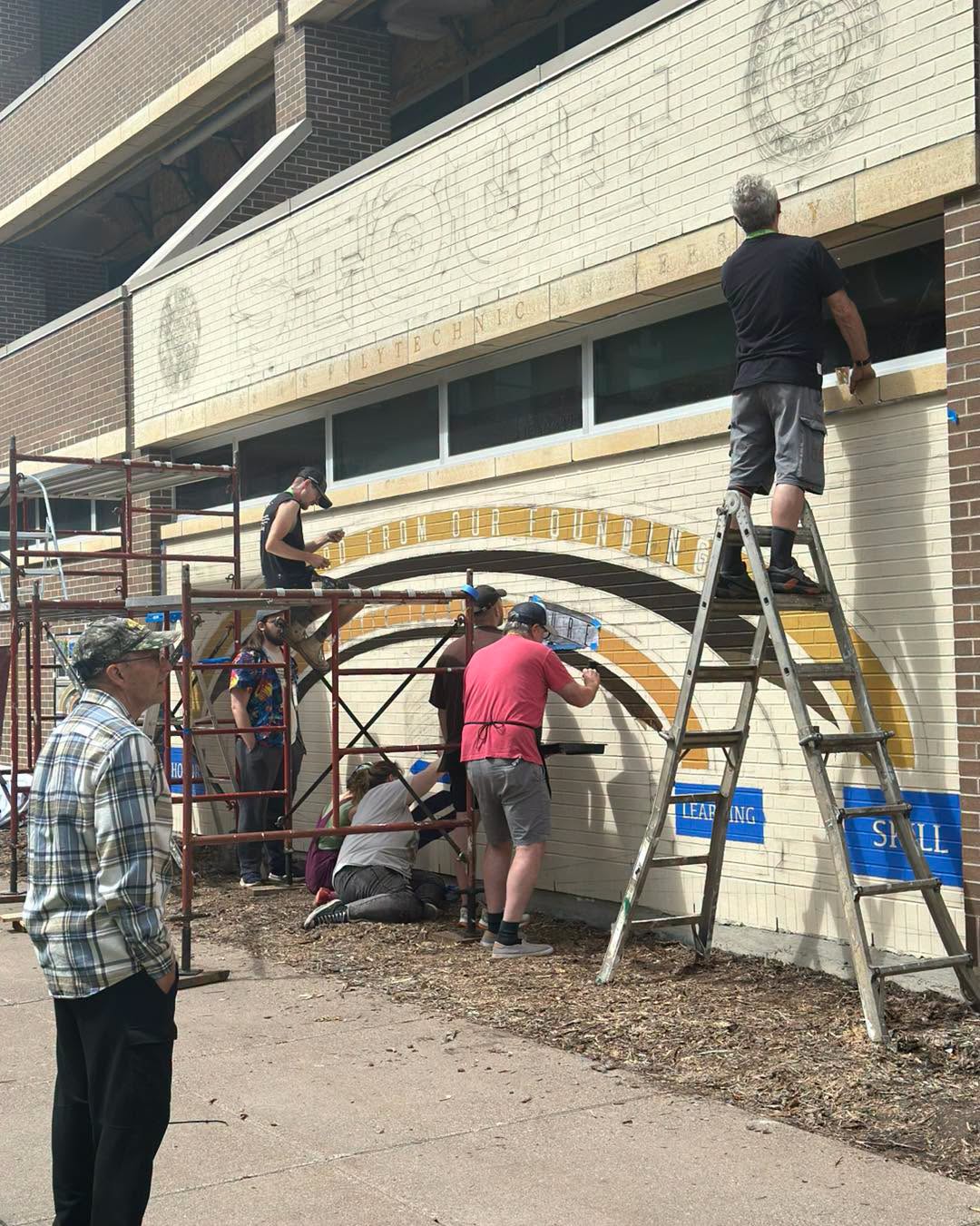 Group of artists painting various sections of a mural with a man looking on