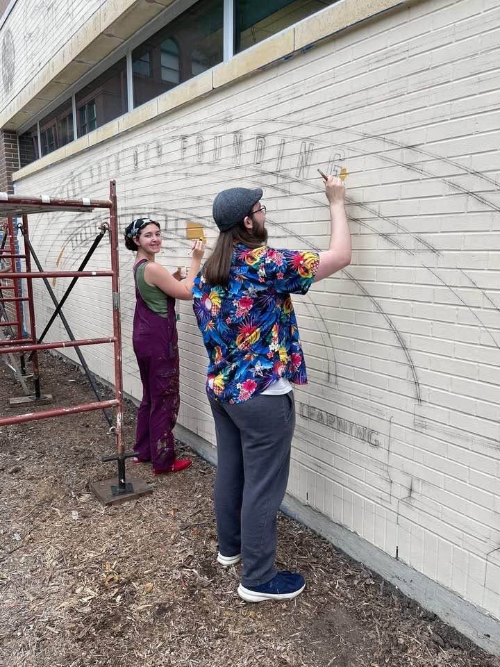 Two artists standing in front of light brick wall stenciling designs