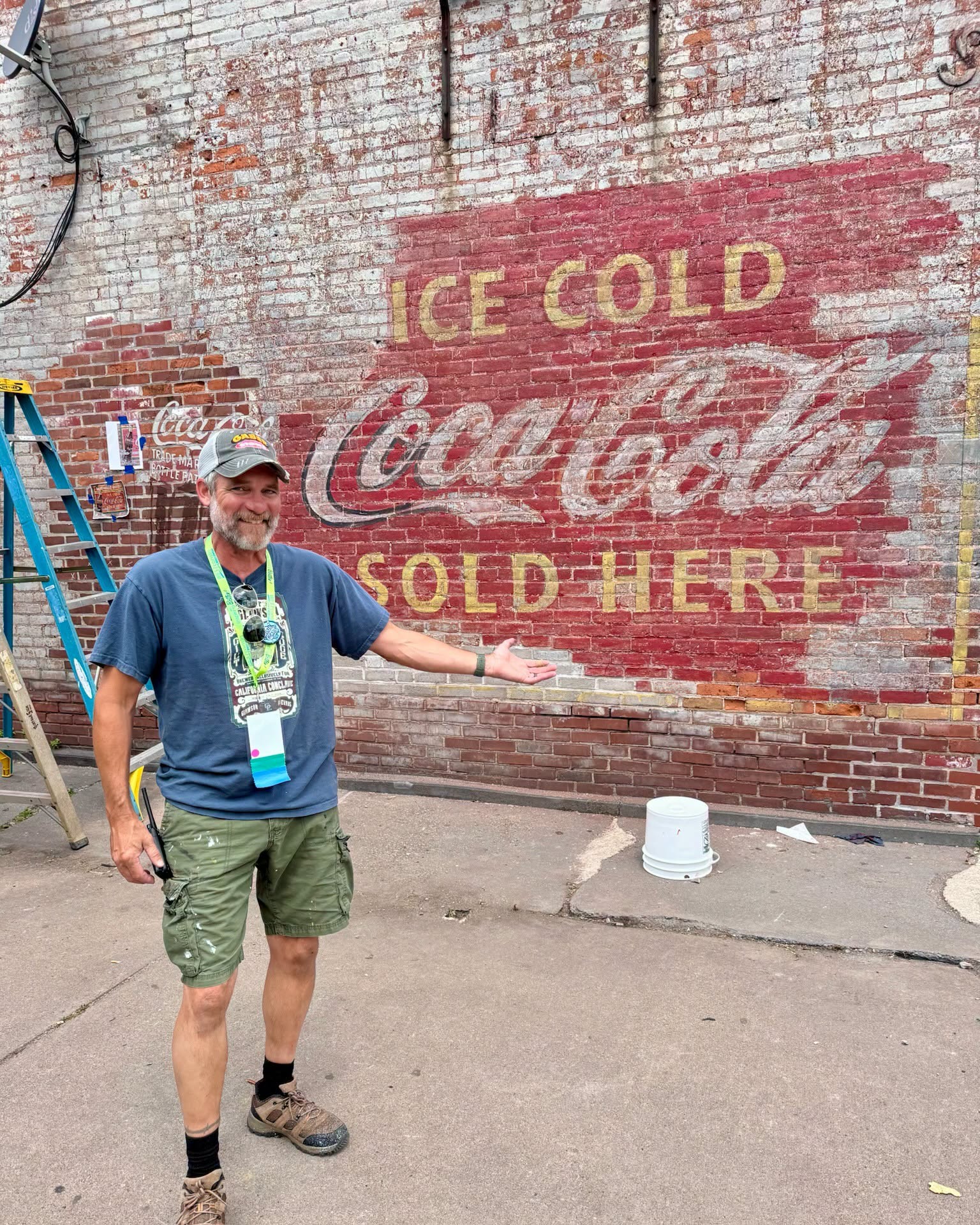 Man holding an arm out to show an old mural painted on and old brick wall saying Ice Cold Coca Cola Sold Here