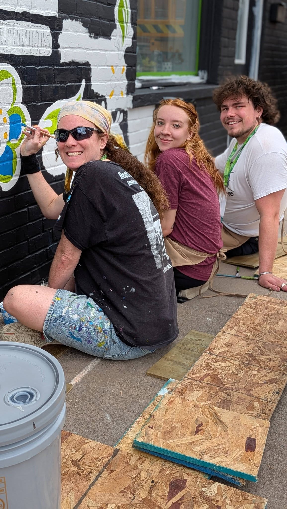 Three people sitting on the ground painting turned to look at the camera