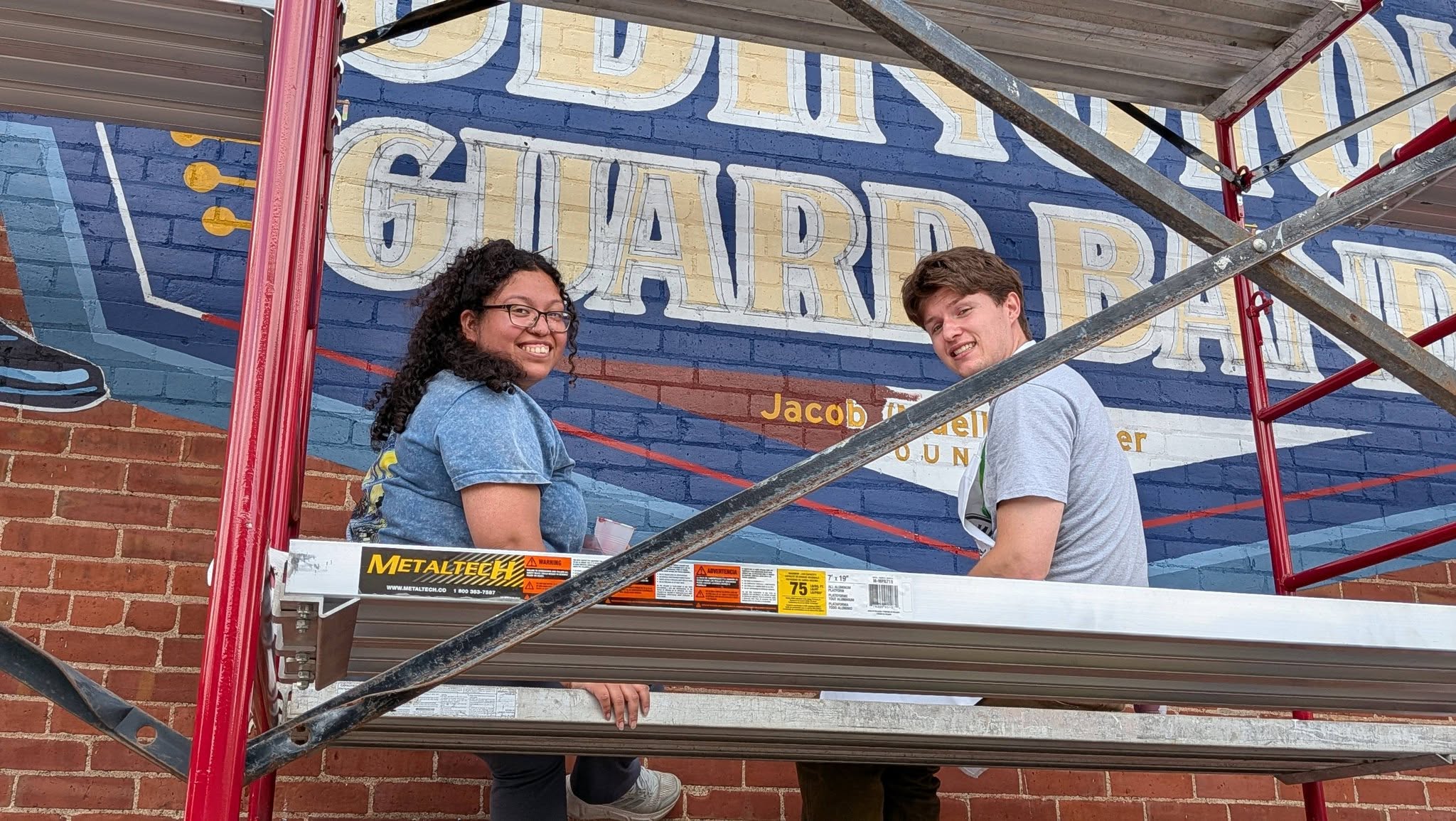 Two people sitting on scaffolding in front of a mural