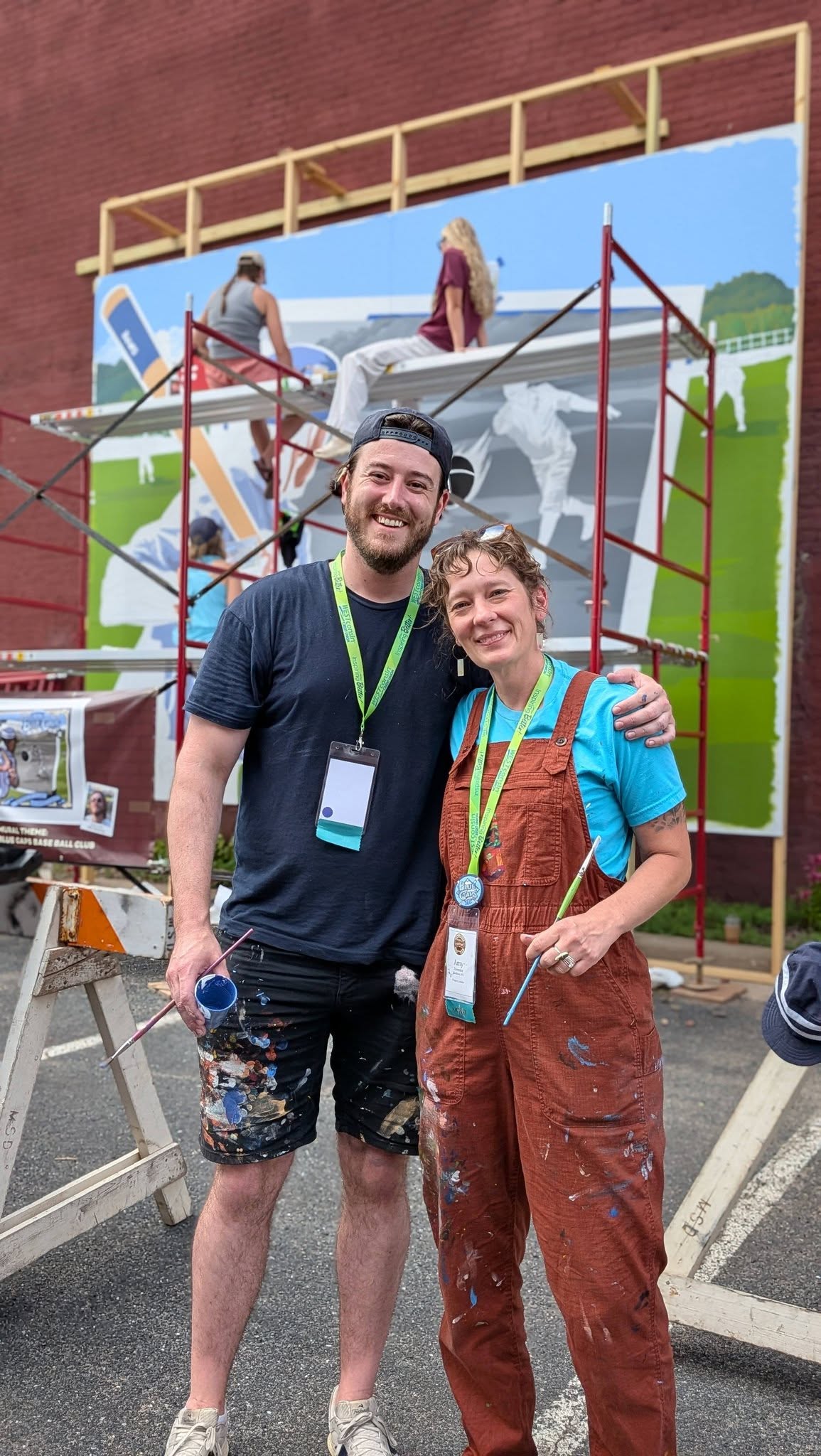 Two artists with paint covered clothing holding paint supplies in front of people on a scaffolding painting a mural