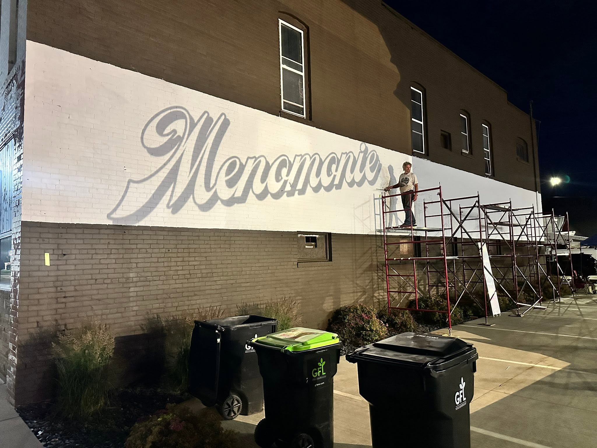 Man standing on scaffolding in front of a wall in the dark with Menomonie projected on the wall