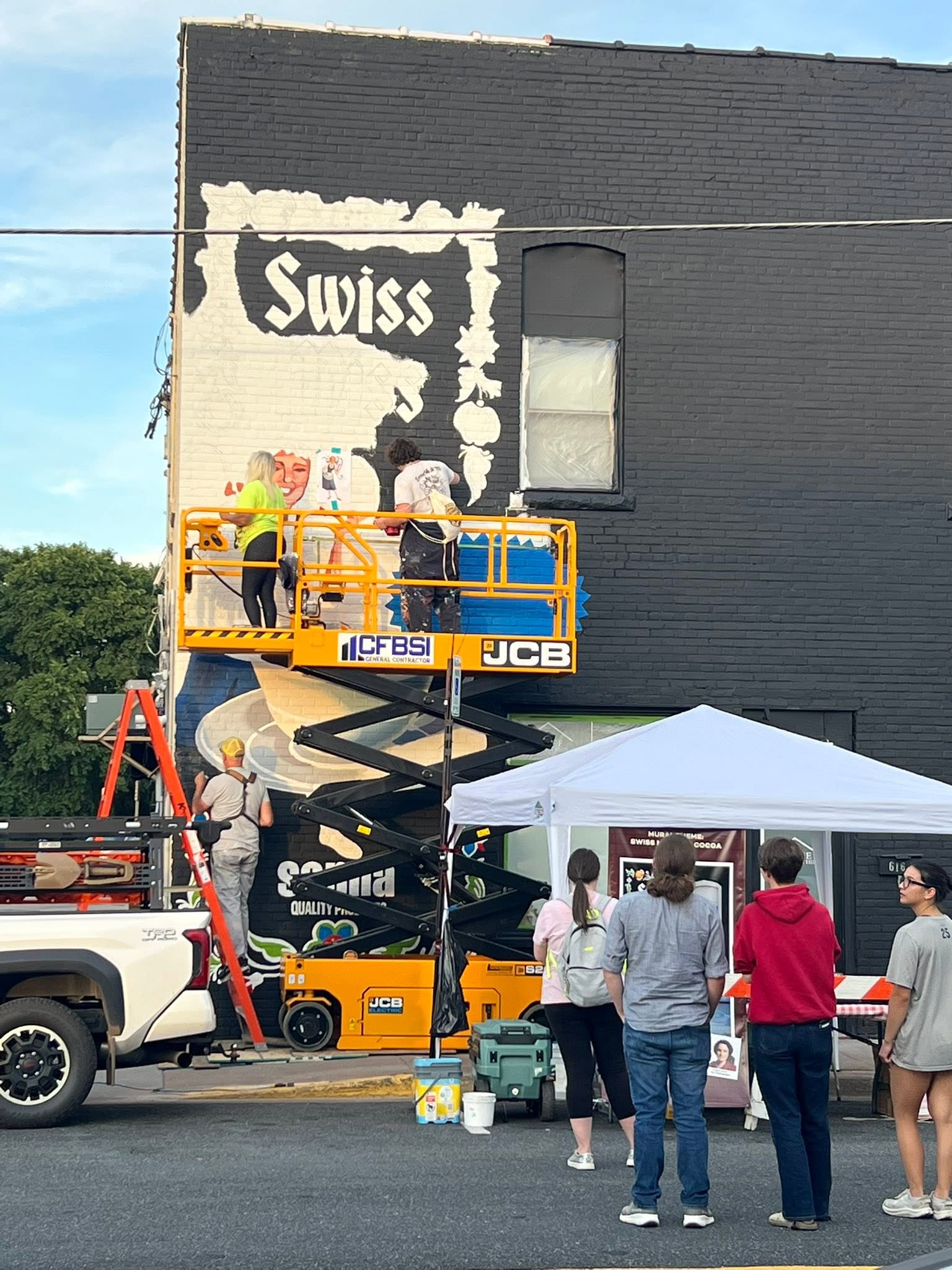 People standing in the street admiring artists in a scissor lift painting a mural