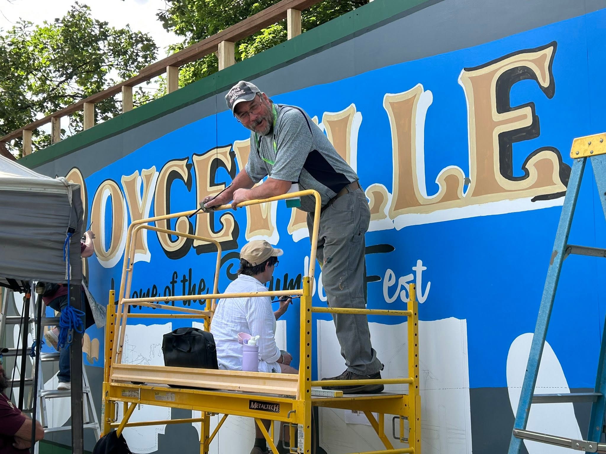 Man on scaffolding looking at the camera in front of a mural getting painted