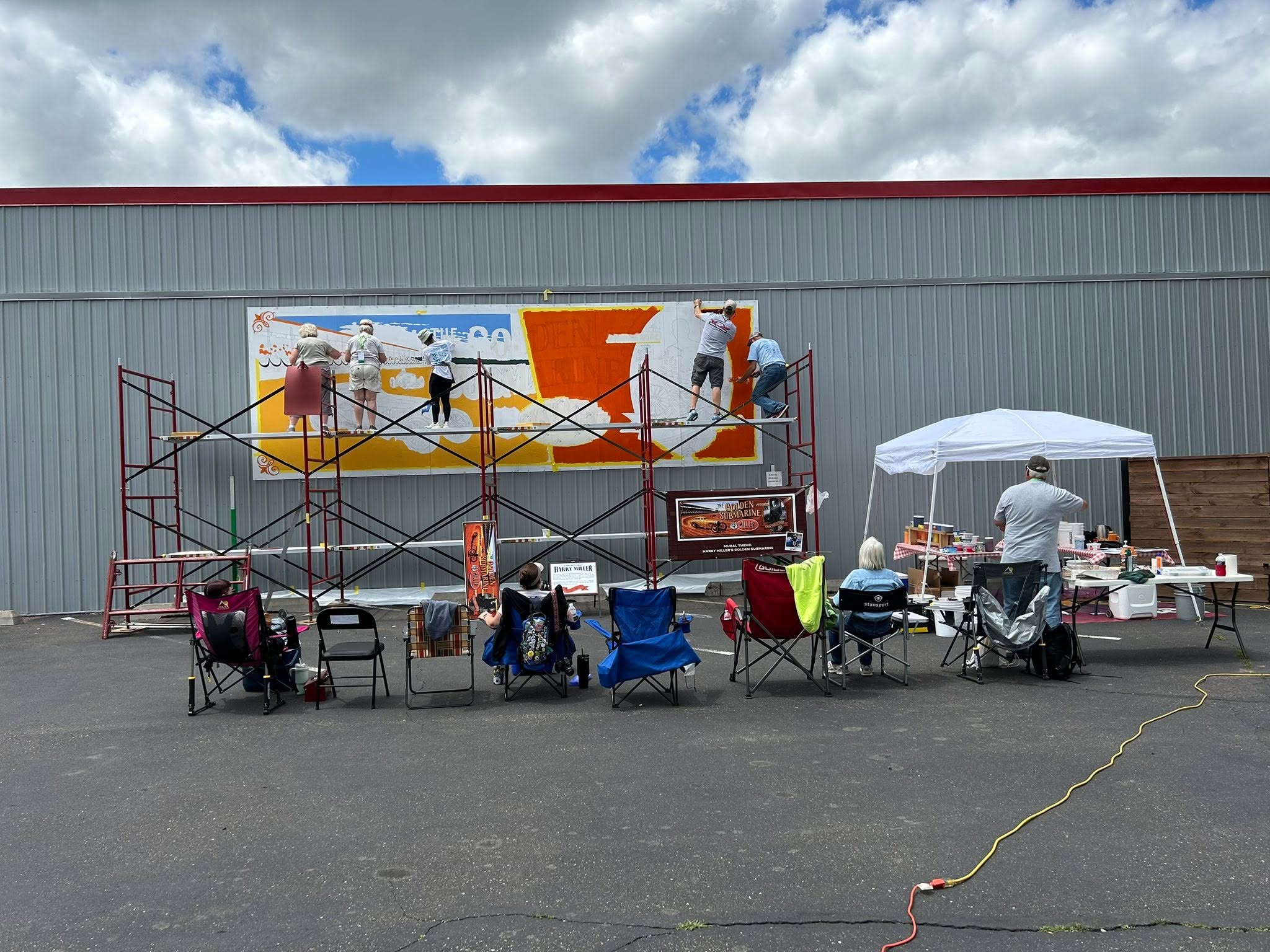 Chairs seated a people watching artists on scaffolding painting a mural