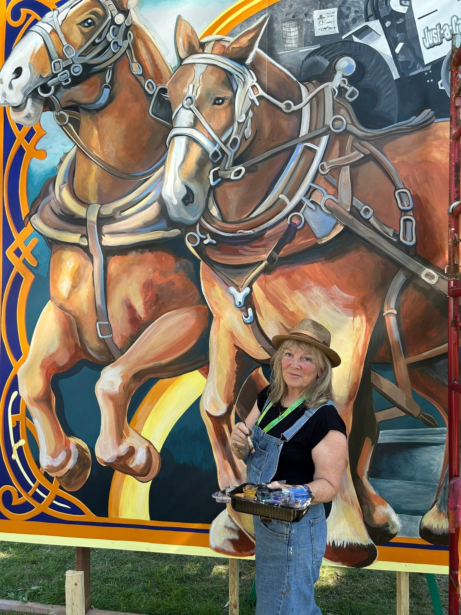 Woman in a hat holding paints and a brush standing in front of painted horses