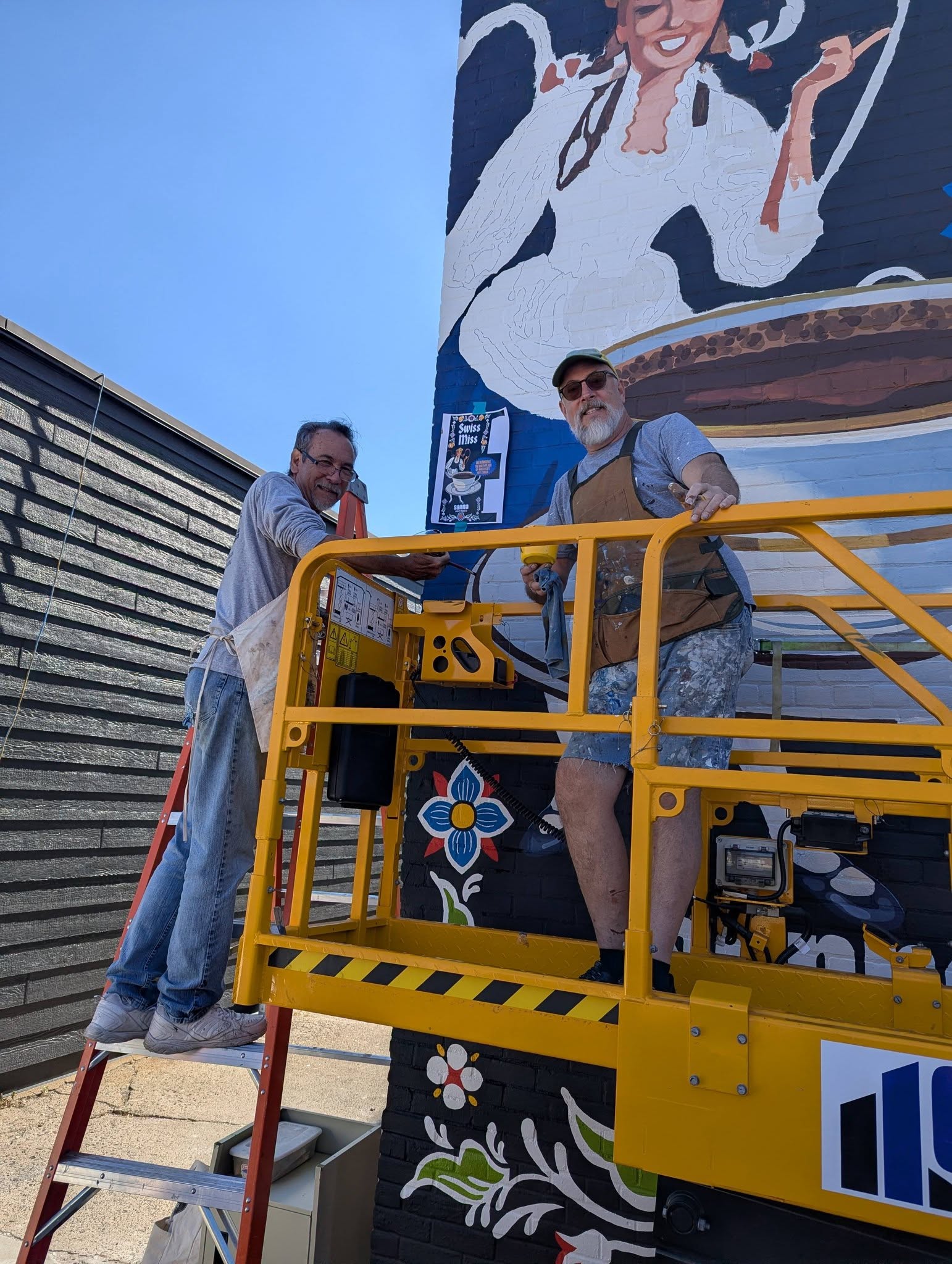A man on a ladder and man in a scissor lift smiling at the camera in front of a mural