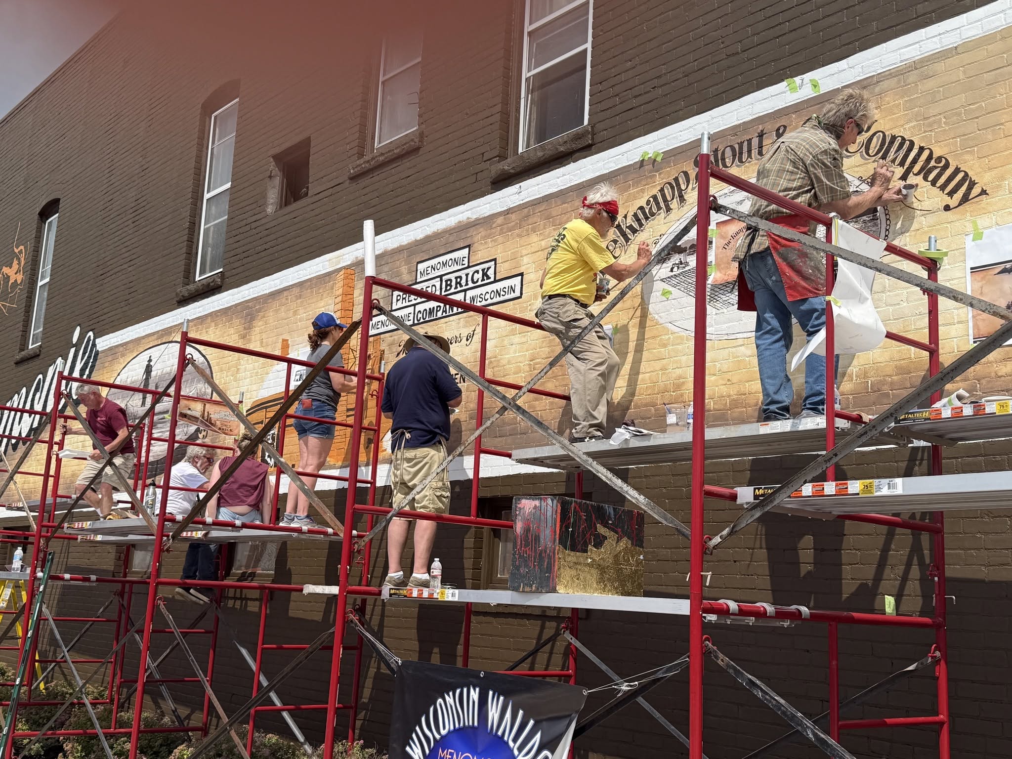 Seven artists sitting and standing on scaffolding painting a mural