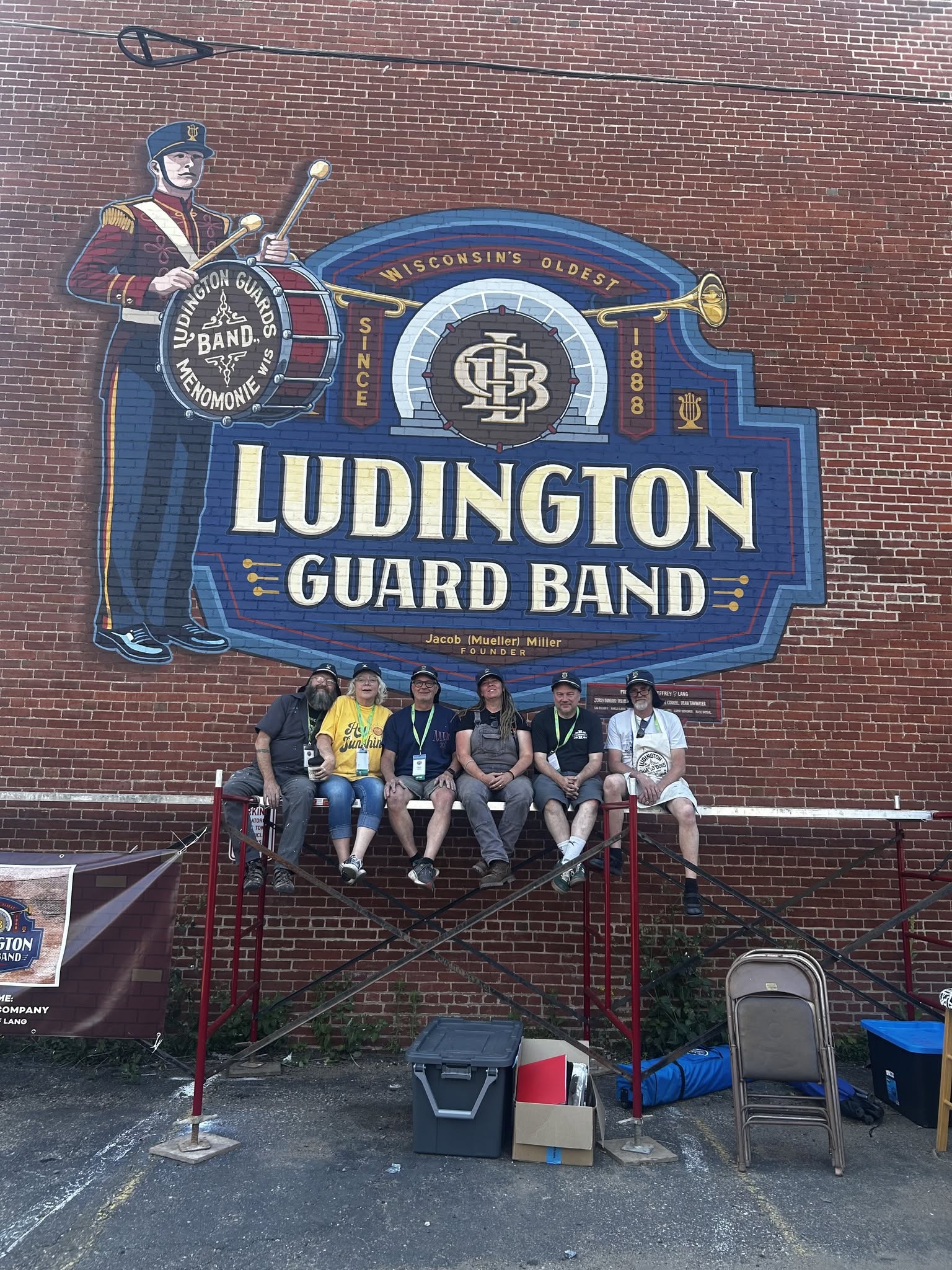 Group of six people sitting on scaffolding in front of the Ludington Guard Band mural