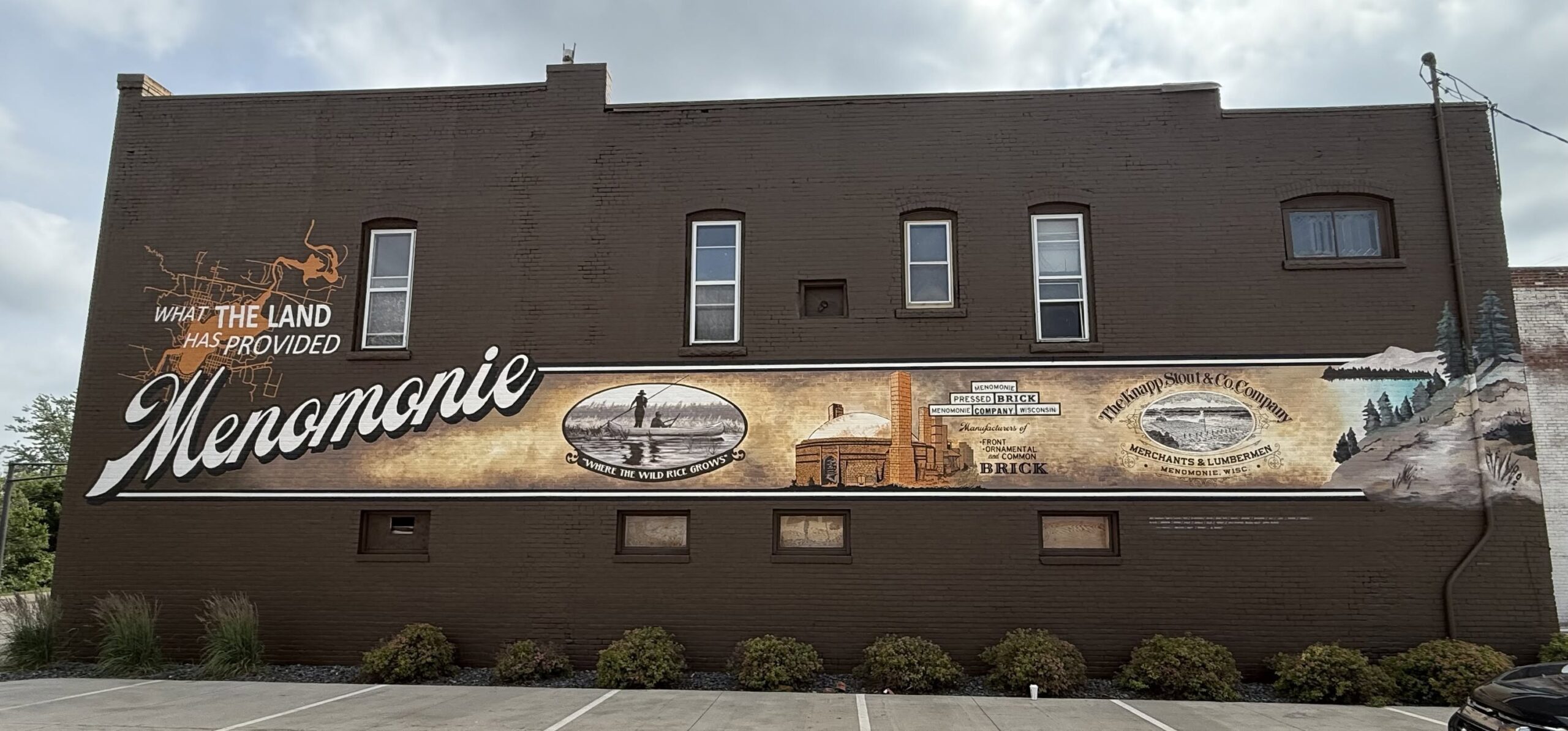 Large long mural painting stretching across a brick wall of What the land has provided Menomonie