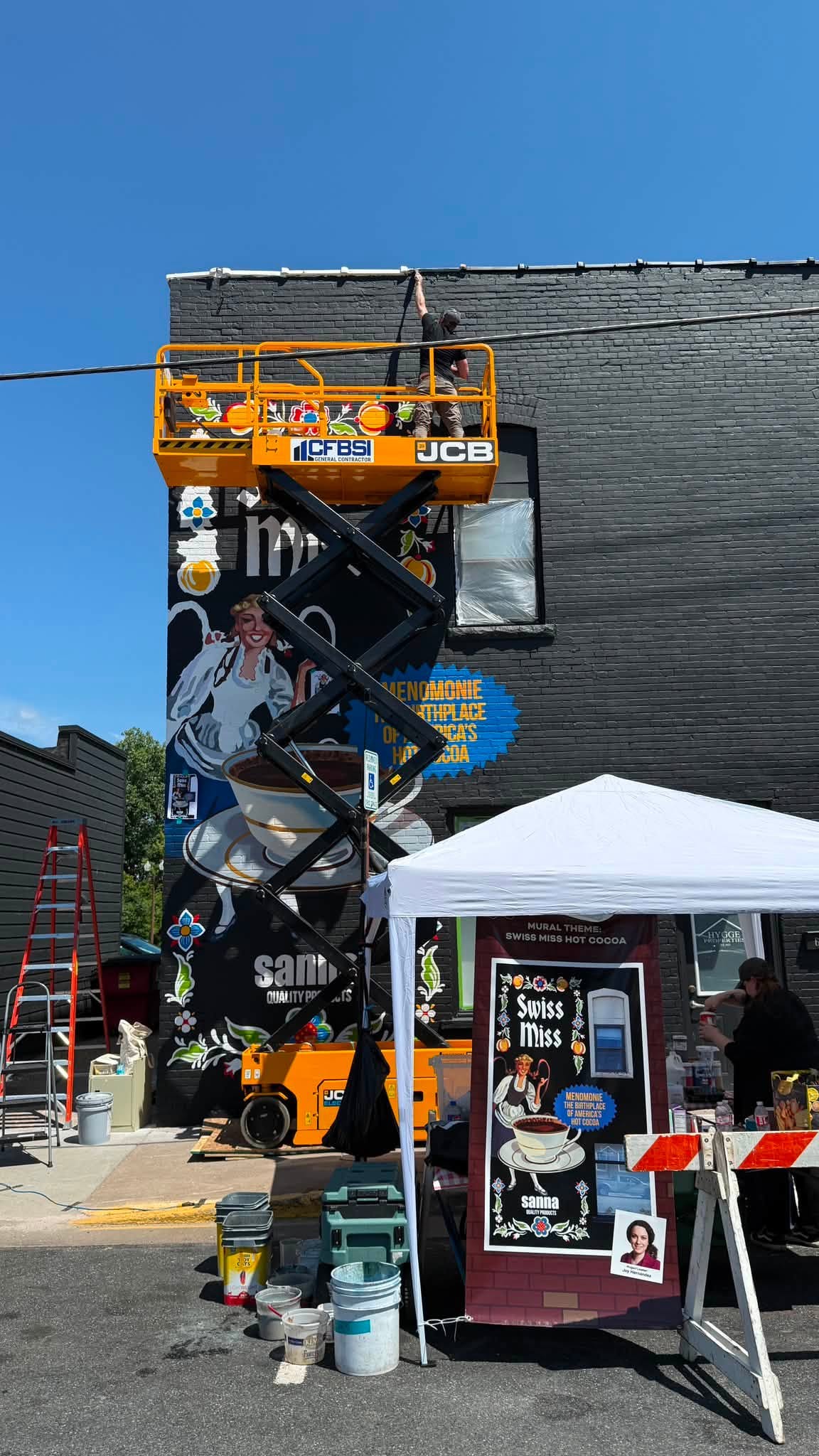 Black and yellow scissor lift extended up in front on a black brick building with a mural