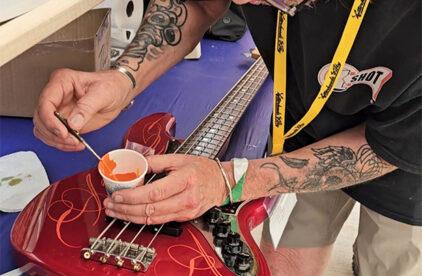 person dipping paintbrush into cup of paint on top of a guitar being painted